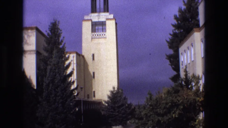 1973 New Mexico home-movie frame with a cream-colored tower, decorative upper detail, evergreen trees, and a side building wing visible at right.