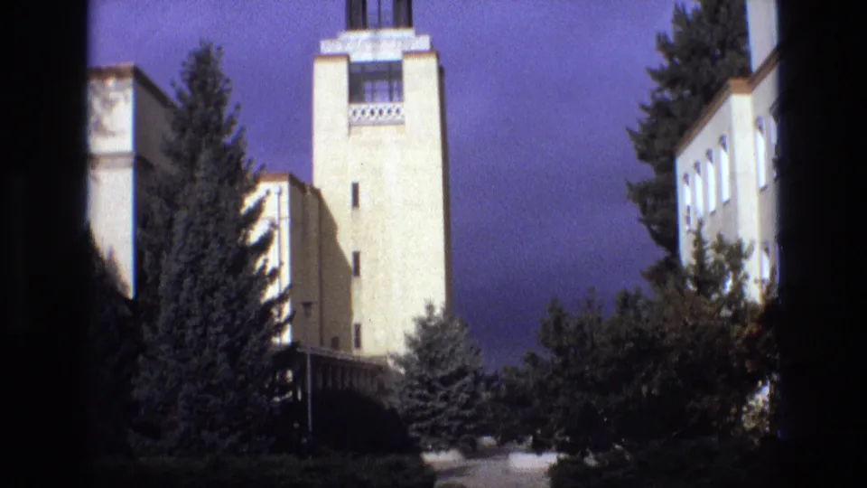 1973 New Mexico home-movie frame showing a pale tower, geometric upper detailing, trees around the building, and dark 8mm frame borders.