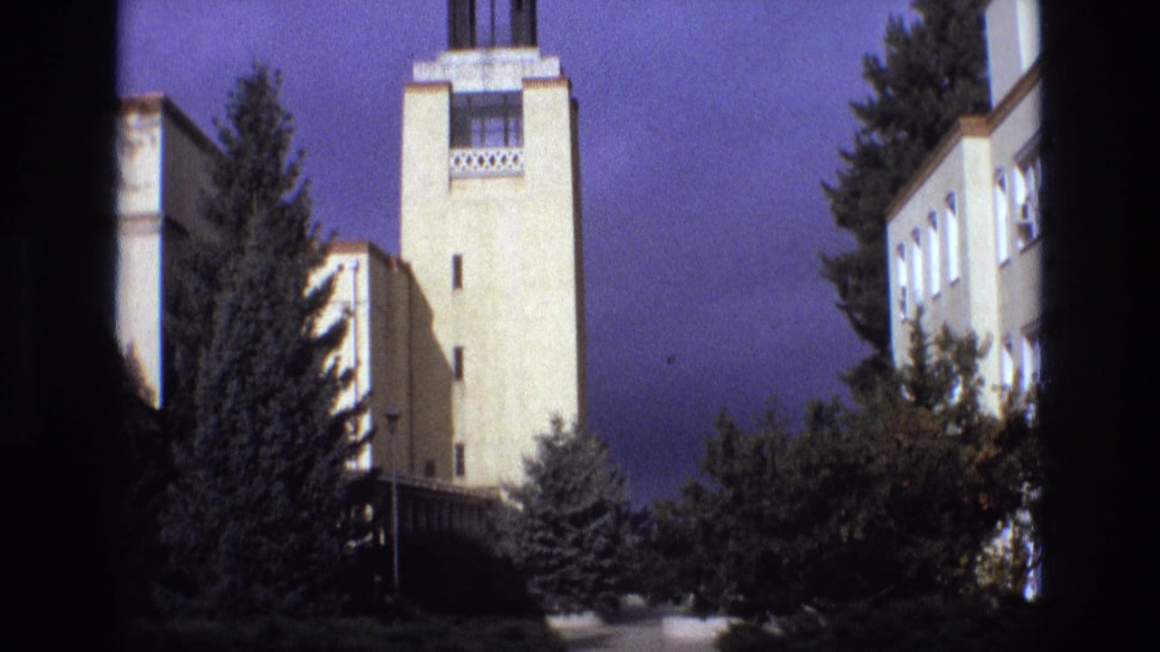 1973 New Mexico 8mm frame showing a pale institutional clock tower or tower facade partly framed by dark vertical edges, with evergreen trees and shrubs in front.