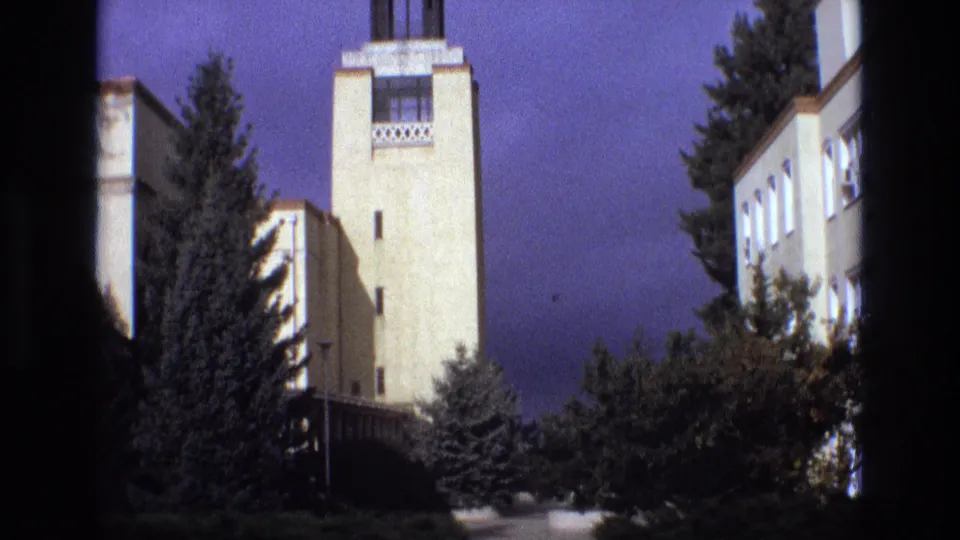 1973 New Mexico 8mm frame showing a pale institutional clock tower or tower facade partly framed by dark vertical edges, with evergreen trees and shrubs in front.