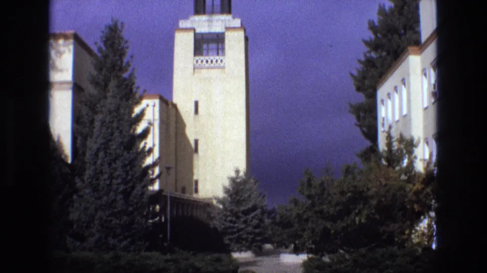 1973 New Mexico 8mm frame of a pale tower beside an institutional building exterior, with evergreens, shrubs, and dark frame edges visible.