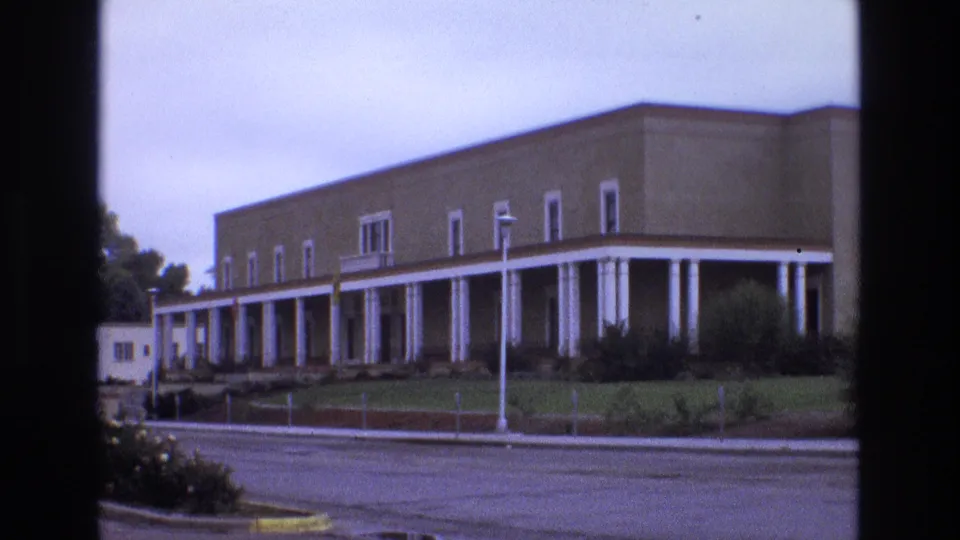 1973 New Mexico 8mm frame showing a brick school-like building with a long white colonnade, lawn, shrubs, street, and streetlamp, seen through dark vertical framing.