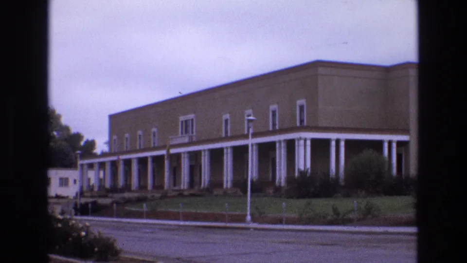 1973 New Mexico 8mm frame of a brick institutional building with a white columned walkway, grassy foreground, shrubs, streetlamp, and paved street.