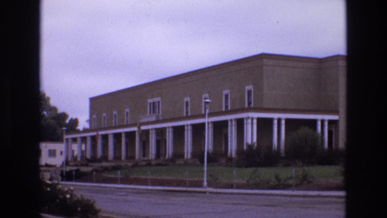 1973 New Mexico 8mm frame showing a long beige stucco-like building with white columns, a covered walkway, landscaping, and a street in the foreground.