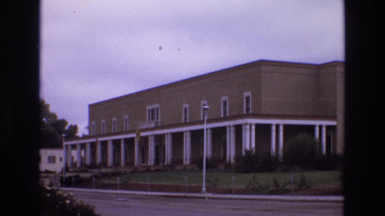 1973 New Mexico frame of a low, flat-roofed building with a long white-columned walkway, seen from across an overcast street.