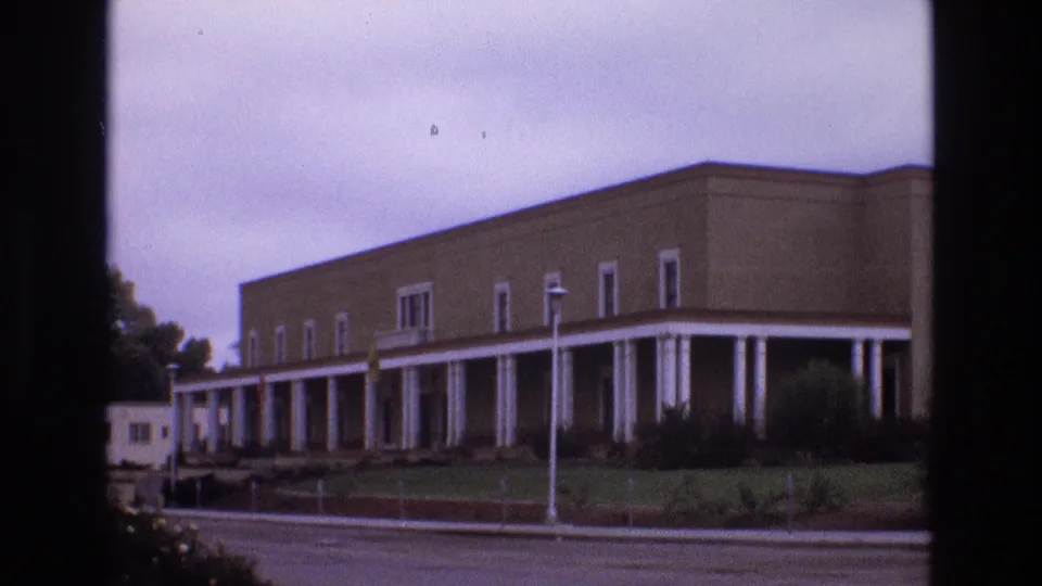 1973 New Mexico frame of a low, flat-roofed building with a long white-columned walkway, seen from across an overcast street.
