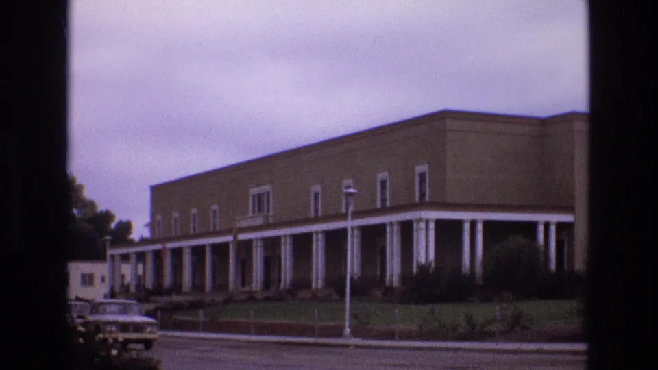 1973 New Mexico home-movie frame with a classic American car moving along the street before a beige building lined with white columns.