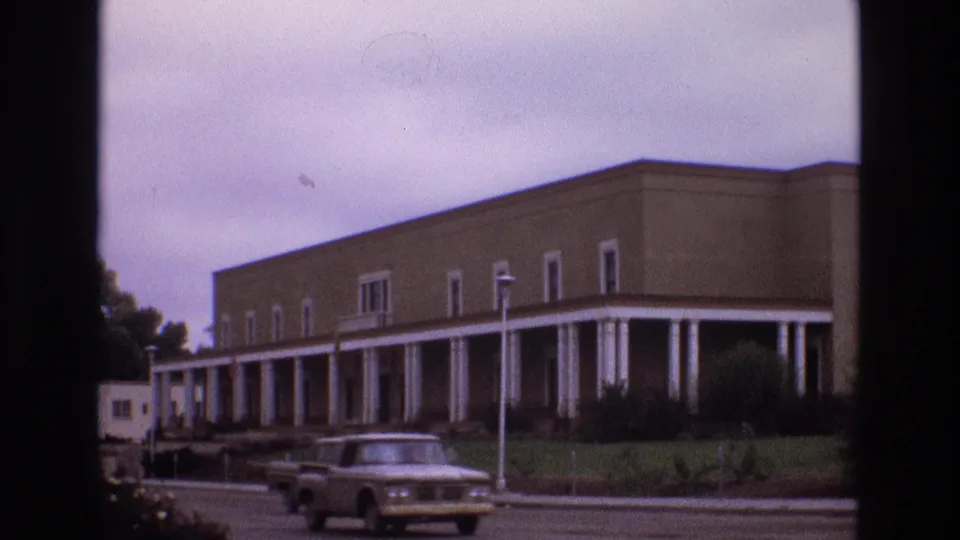 1973 New Mexico 8mm frame showing a classic car crossing the foreground while a long beige building with white columns remains in the background.