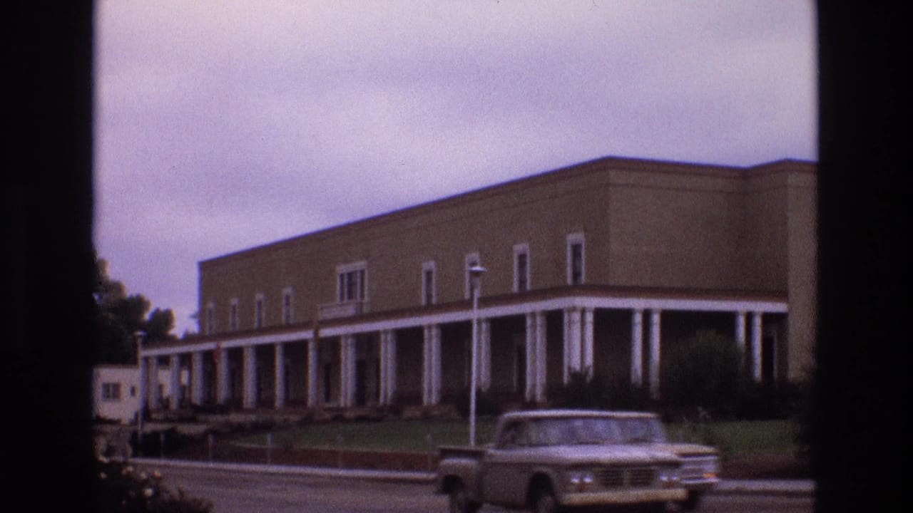 1973 New Mexico 8mm frame showing a large tan building with white columns, landscaped grounds, and a pickup truck moving along the road in front.