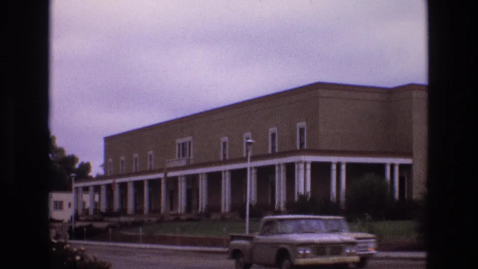 1973 New Mexico 8mm frame showing a large tan building with white columns, landscaped grounds, and a pickup truck moving along the road in front.