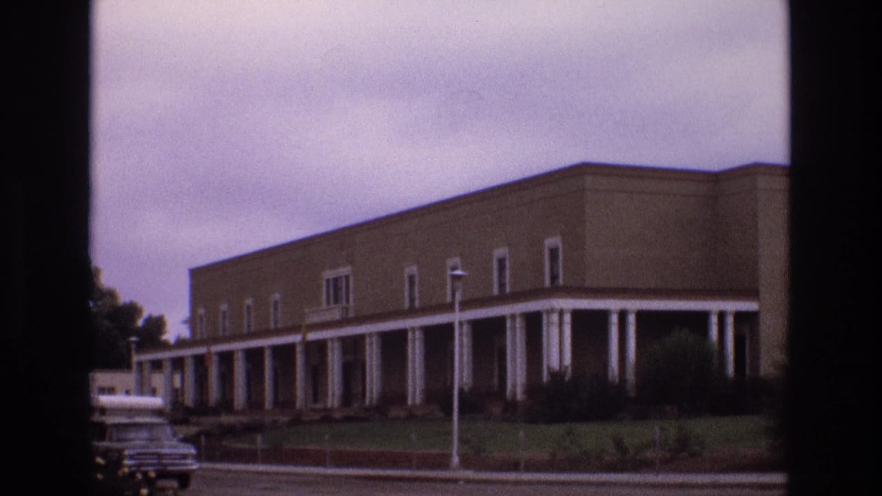 1973 New Mexico exterior view of a large tan columned building with a curved facade, manicured grass, shrubs, and a vehicle partly visible near the road.
