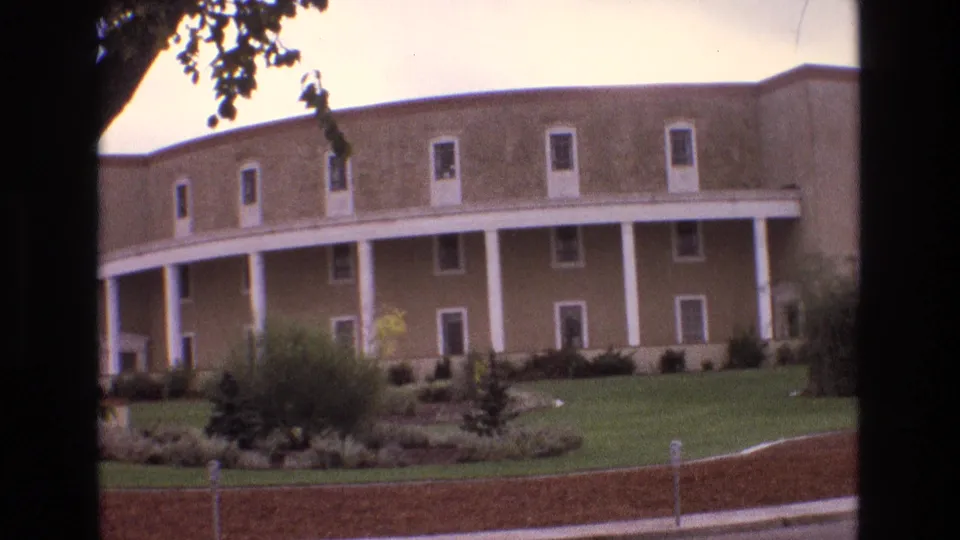 1973 New Mexico 8mm frame showing a closer roadside view of the landscaped grounds and white-columned facade of a large tan building.