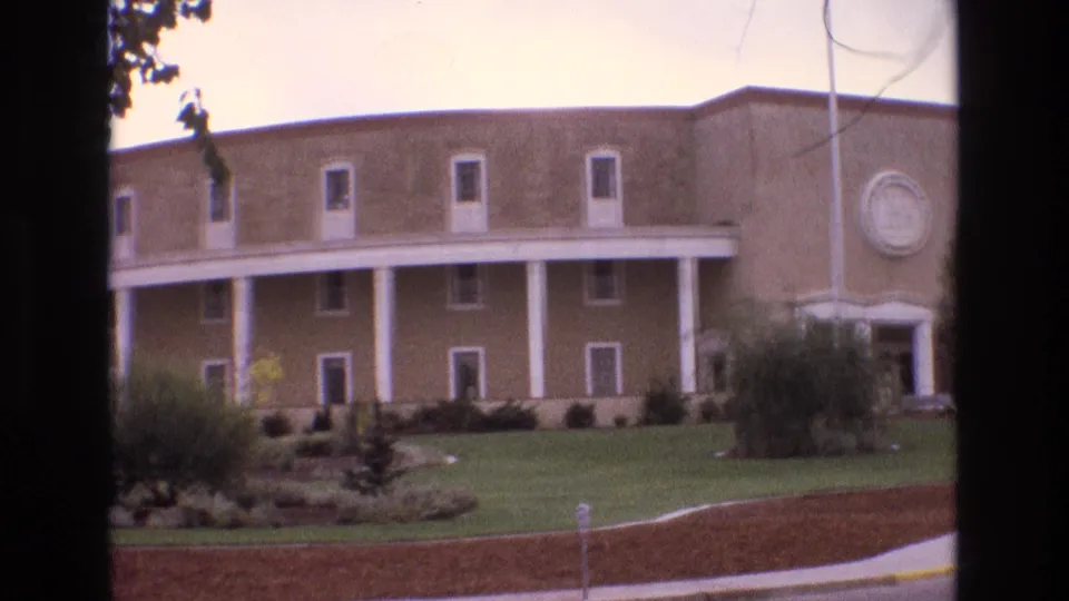 1973 New Mexico exterior frame of the tan columned building, showing landscaped grounds, a covered porch, and a circular decorative feature on the right wall.