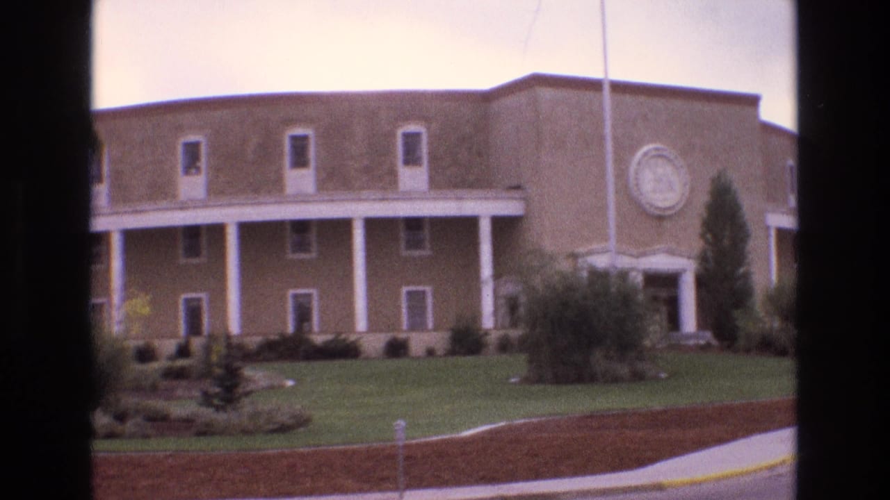 1973 New Mexico 8mm frame showing a large curved tan building with white columns, a circular wall emblem, landscaped lawn, shrubs, and a roadway in the foreground.