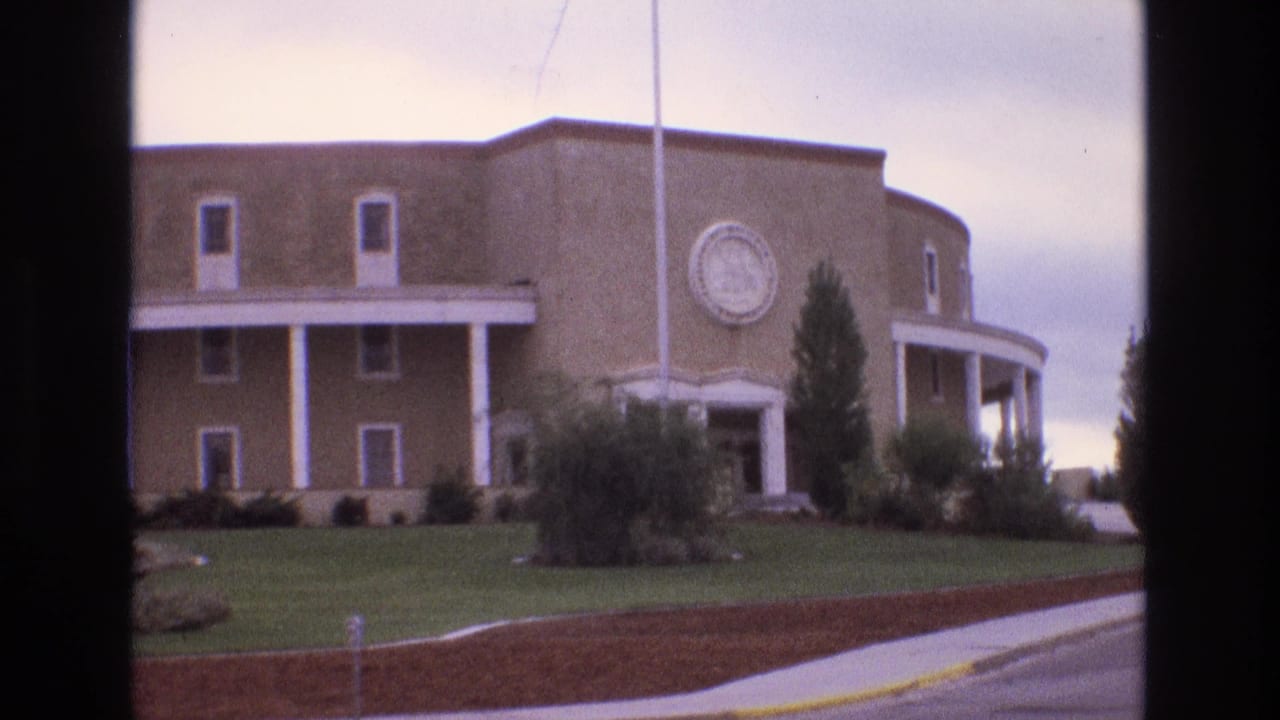 1973 New Mexico 8mm frame with the tan curved building centered behind a lawn, white columns, circular emblem, shrubs, trees, sidewalk, and curb visible from across the road.
