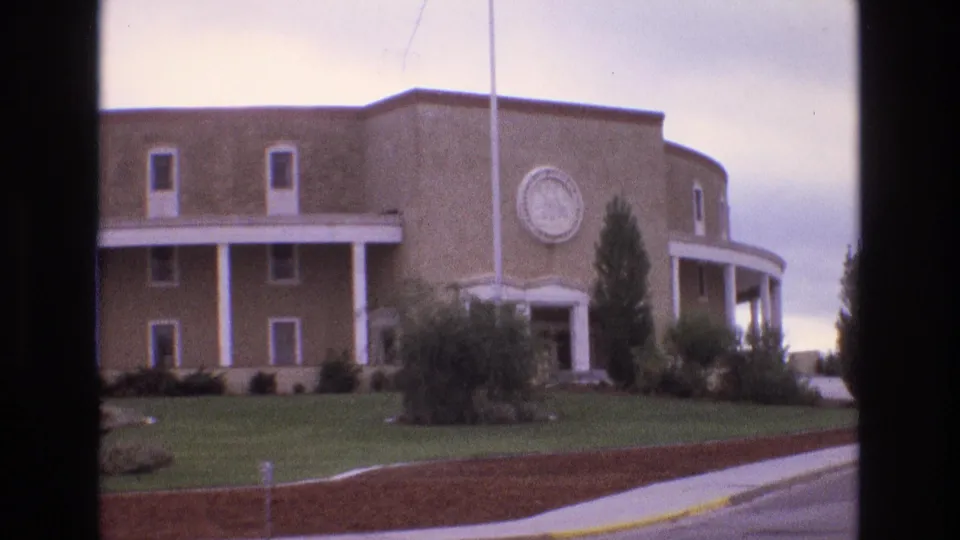 1973 New Mexico 8mm frame with the tan curved building centered behind a lawn, white columns, circular emblem, shrubs, trees, sidewalk, and curb visible from across the road.
