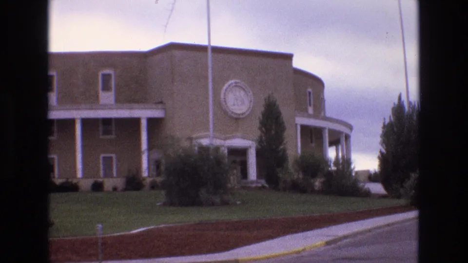 1973 New Mexico 8mm frame showing an off-center view of the tan building, circular emblem, white columned porch, shrubs, lawn, roadway, and dark vertical film border.