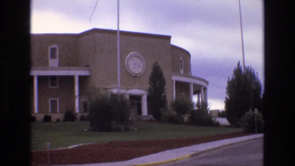 1973 New Mexico 8mm frame showing the curved tan building with a circular emblem, white columns, landscaped lawn, shrubs, trees, roadway, and cloudy sky.