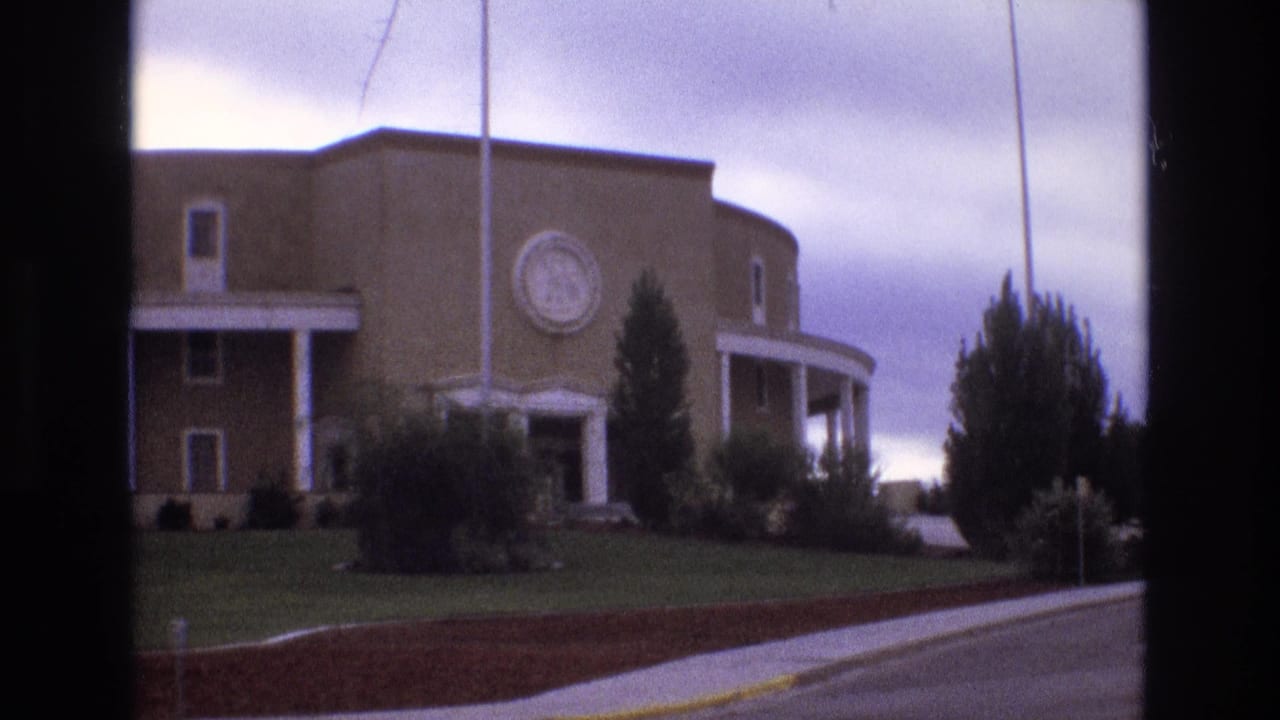 1973 New Mexico 8mm frame showing a brick civic-style building with rounded lines, white columns, a circular emblem, flagpoles, landscaped lawn, shrubs, and a street edge.