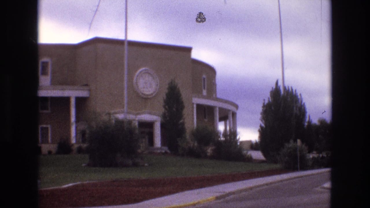 Similar view of the brick building facade, circular emblem, flagpoles, shrubs, lawn, and roadway.