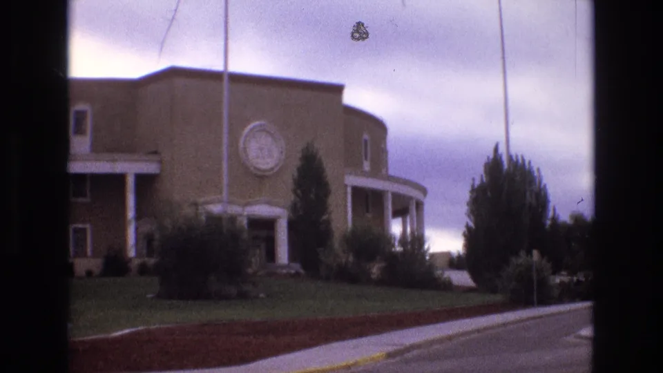 1973 New Mexico 8mm frame holding on the brick institutional facade with a round emblem, white-columned entrance areas, flagpoles, manicured lawn, shrubs, and adjacent road.