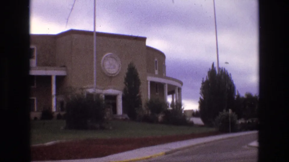 1973 New Mexico 8mm frame with a closer handheld view of the brick building, circular emblem, white columns, flagpoles, planted lawn, shrubs, street curb, and cloudy sky.
