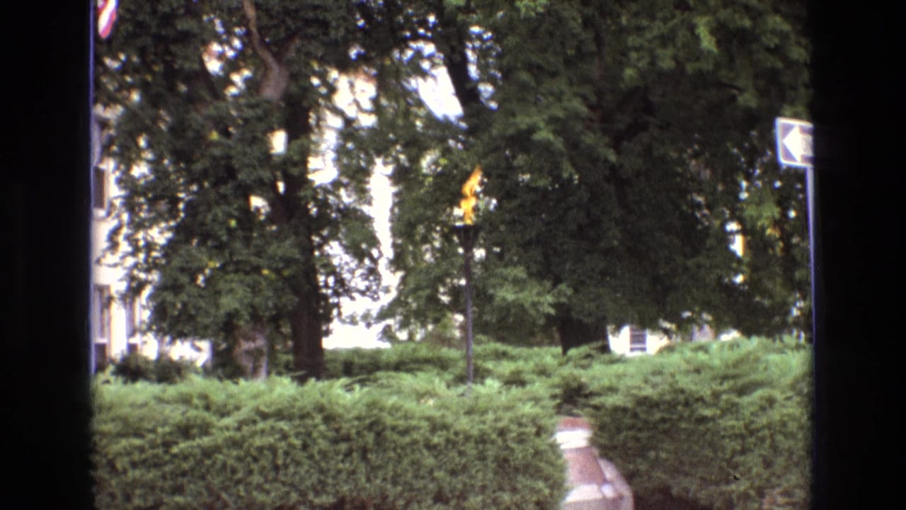 1973 New Mexico 8mm frame showing a small gas flame burning from the top of a pole in a landscaped outdoor area with hedges, trees, a path, and a white arrow sign.