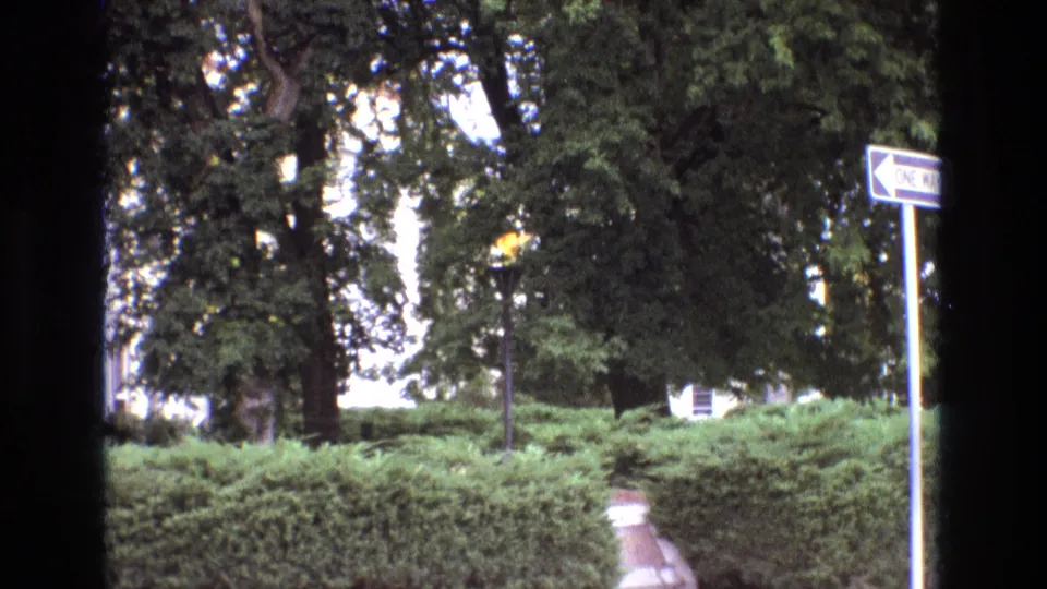 1973 New Mexico 8mm frame showing a small gas flame above a pole in a landscaped outdoor area, with hedges in the foreground, trees behind, a path, and a white arrow sign at right.