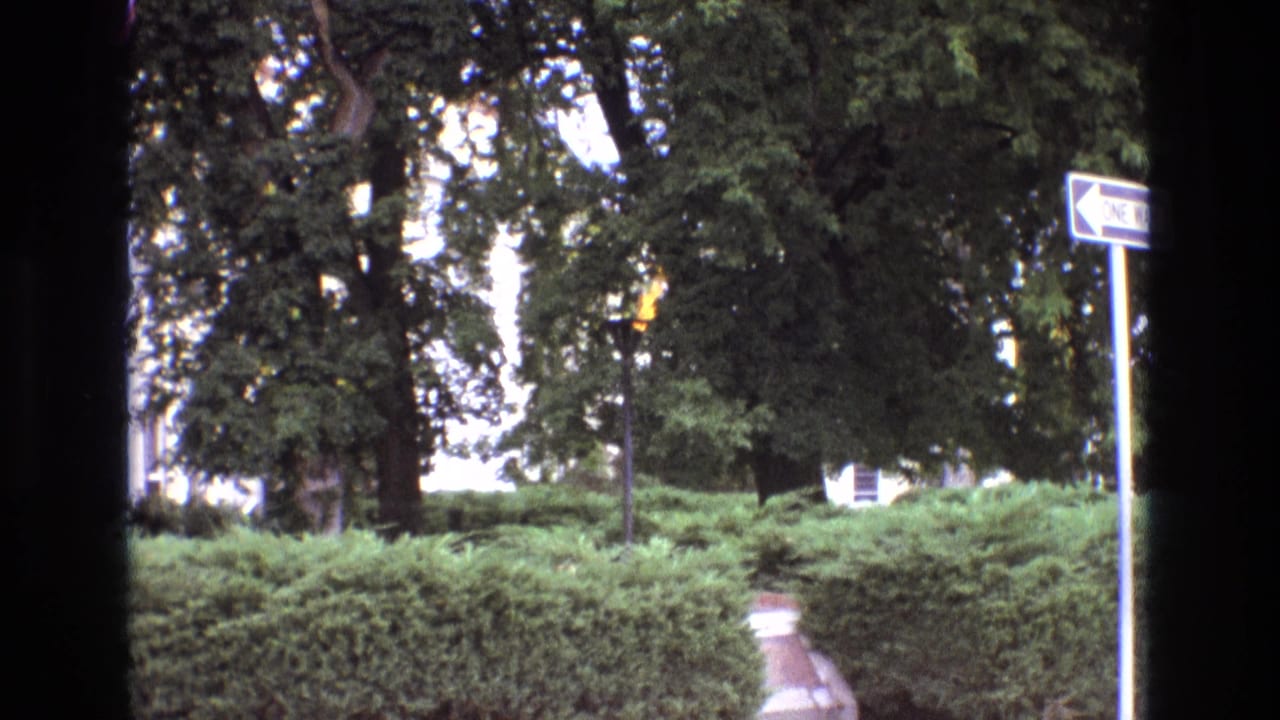 1973 New Mexico 8mm frame showing a landscaped park or roadside area with green hedges, mature trees, a walkway, a one-way sign, and heavy dark borders from the filming position.