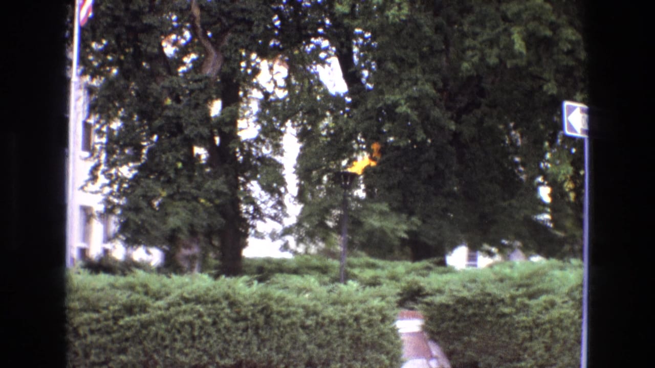 1973 New Mexico 8mm frame showing a small torch-like flame near manicured hedges, a paved walkway, mature trees, and the edge of a building facade.