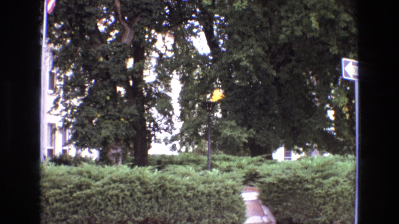 1973 New Mexico 8mm frame with a torch-like flame set against landscaped hedges, tall trees, a walkway, and a partially visible exterior wall.