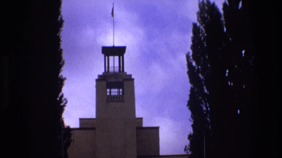 1973 New Mexico 8mm frame looking upward at a towered building with a flagpole, framed by tall trees against a purple-tinged sky.