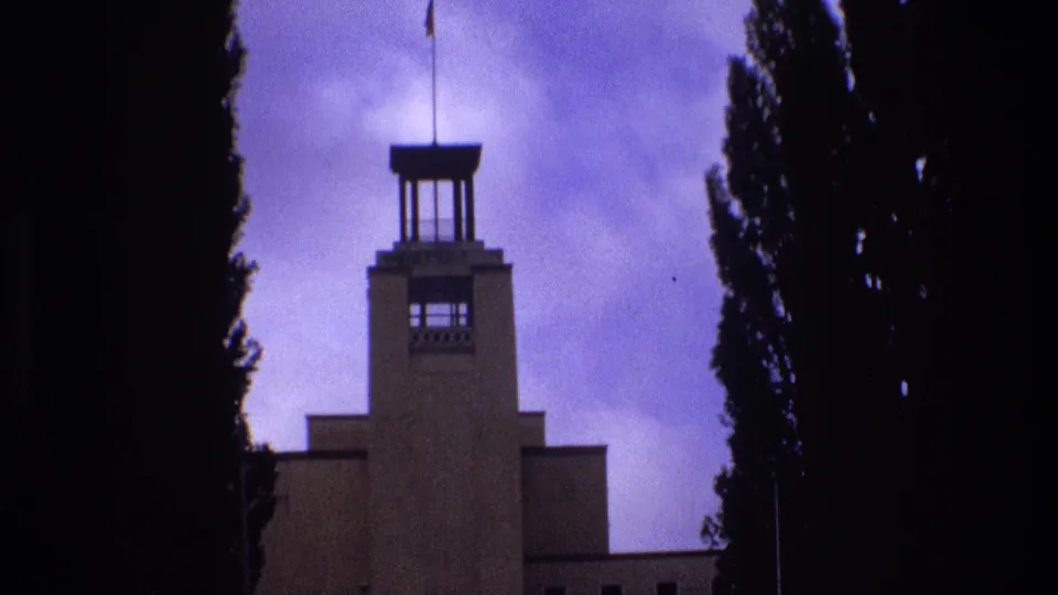 1973 New Mexico 8mm frame showing a towered building with a flagpole and flag, bordered by tall dark trees beneath a purple-shifted sky.