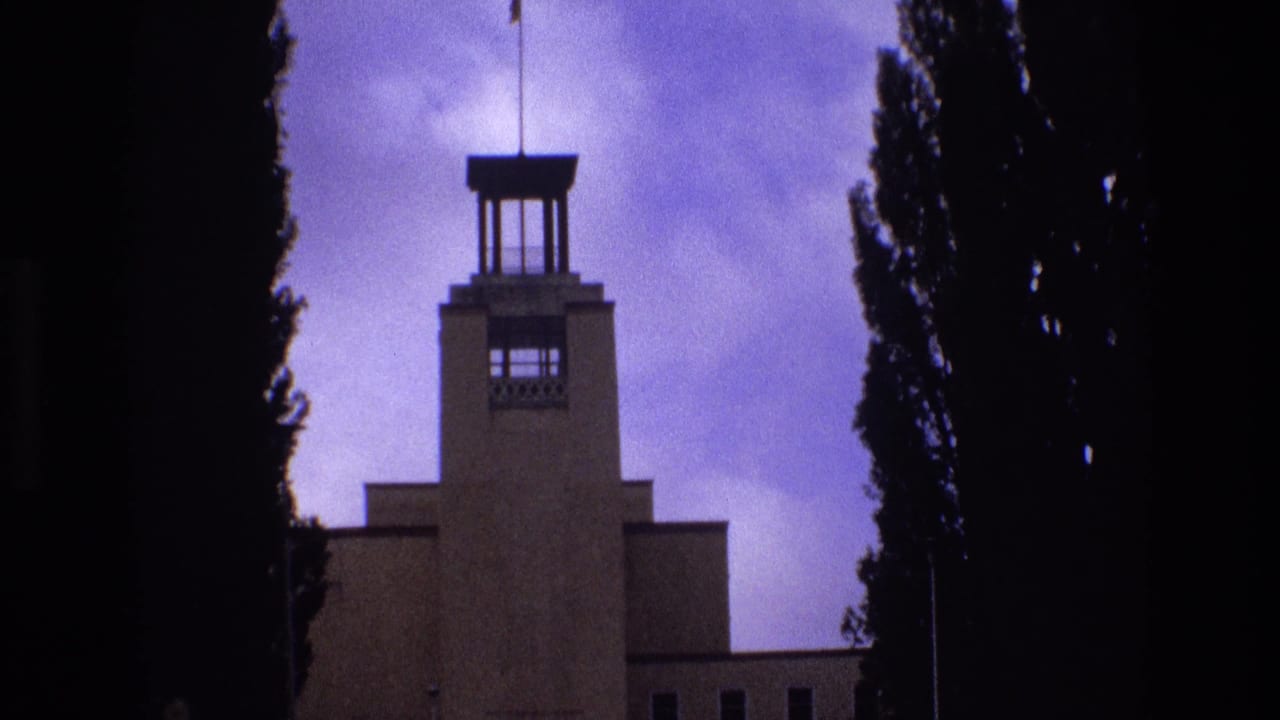 1973 New Mexico 8mm frame showing a stepped towered building with tall upper openings, partly obscured by dark tree silhouettes and a cloudy purple-blue sky.