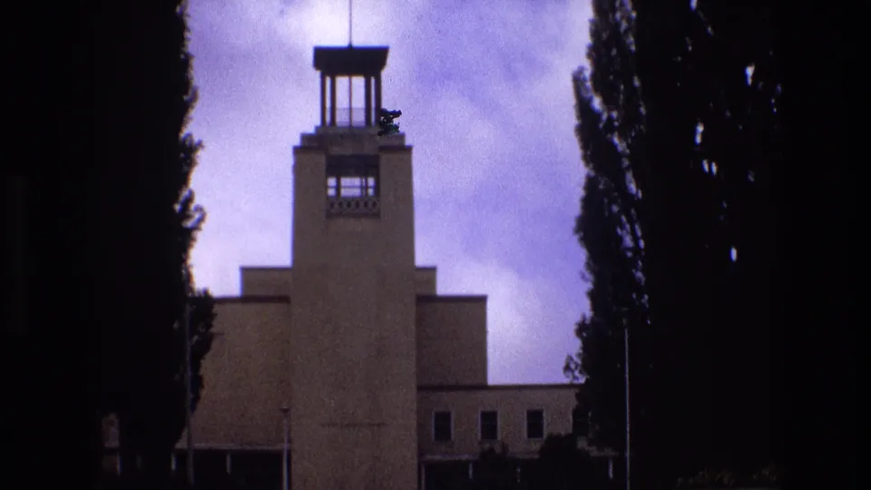 1973 New Mexico 8mm frame showing the towered facade from a slightly lower angle, with a long lower wing, dark tree borders, and faint landscaping at the base.