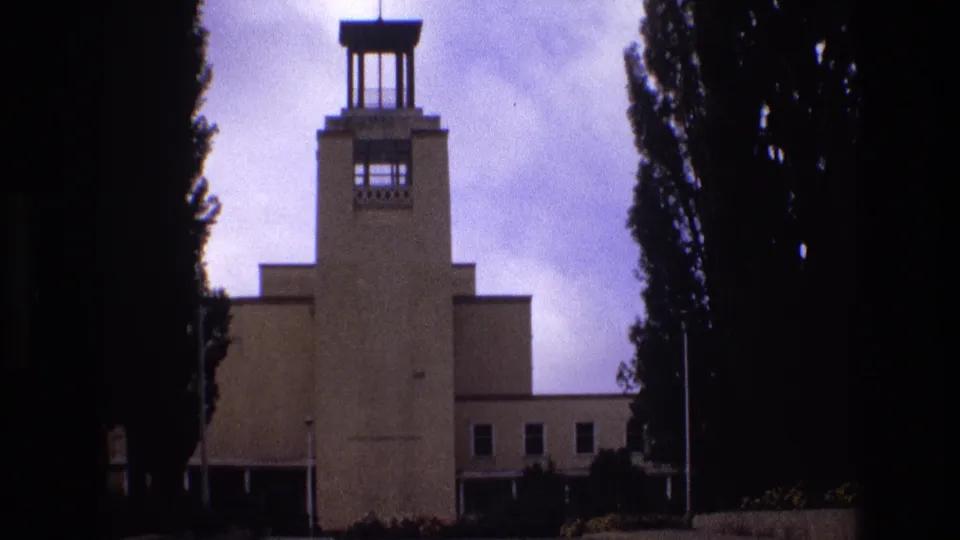 1973 New Mexico 8mm frame showing the towered exterior, side windows, a narrow pole in the foreground, dark trees, and a purple-blue overcast sky.