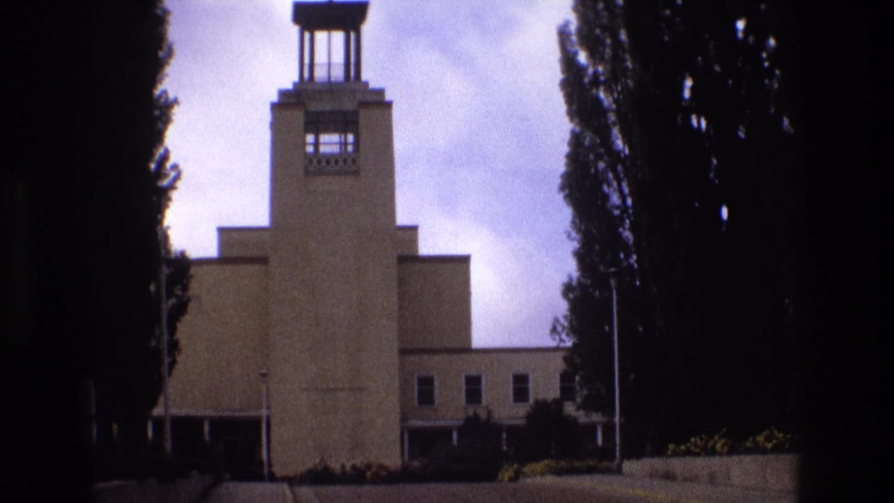 1973 New Mexico 8mm frame showing a tan Art Deco-influenced building with a central tower, flagpole, paved approach, landscaping, and tall trees at the sides.