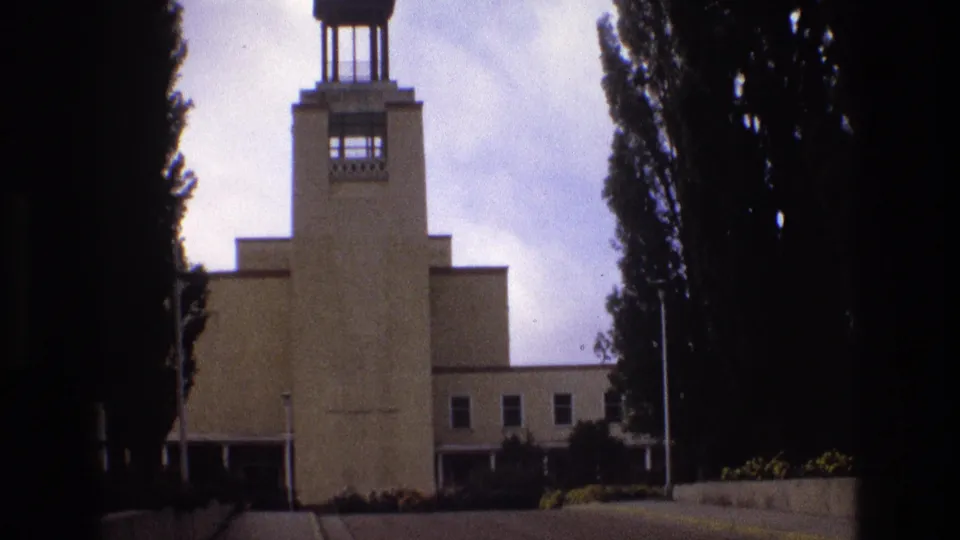 1973 New Mexico 8mm frame showing the building's tower and stepped massing from a slightly shifted handheld angle, with trees, walkway, and landscaping visible.