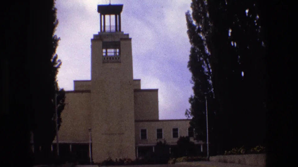 Final 1973 New Mexico frame of the tan institutional-looking exterior, with the central tower, rectangular windows, flagpole, dark trees, and faded 8mm color visible.