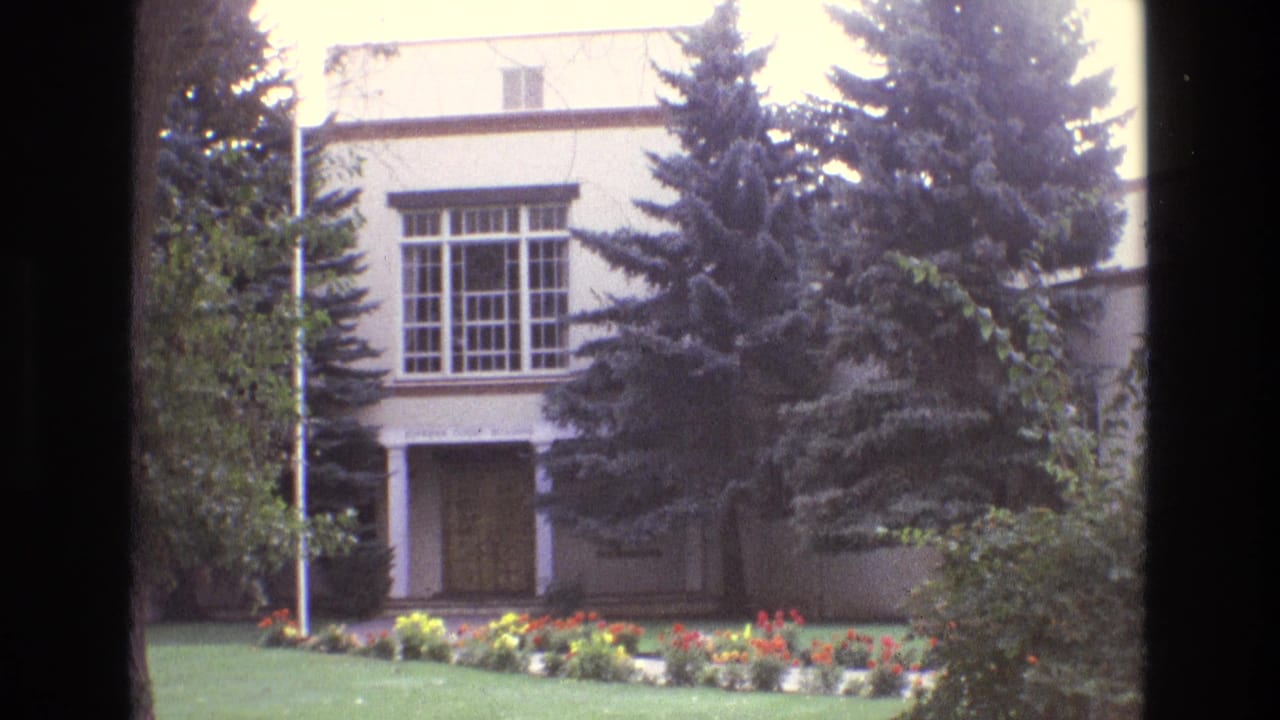 1973 New Mexico 8mm frame showing a white two-story building with a large gridded window, covered entrance, evergreens, lawn, and flowers.