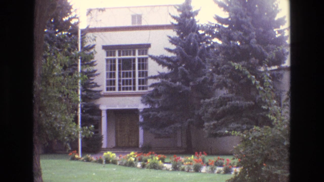 1973 New Mexico 8mm frame of a white building facade with a multi-pane window, recessed doorway, tall evergreens, shrubs, lawn, and flower bed.