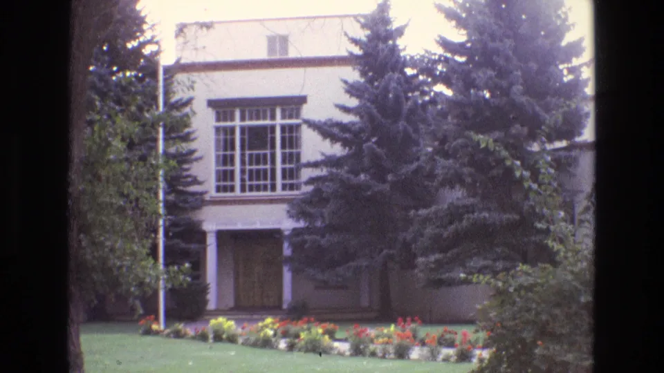 1973 New Mexico home-movie frame showing the same white building exterior with evergreens, a covered entrance, bright lawn, and low flower bed.