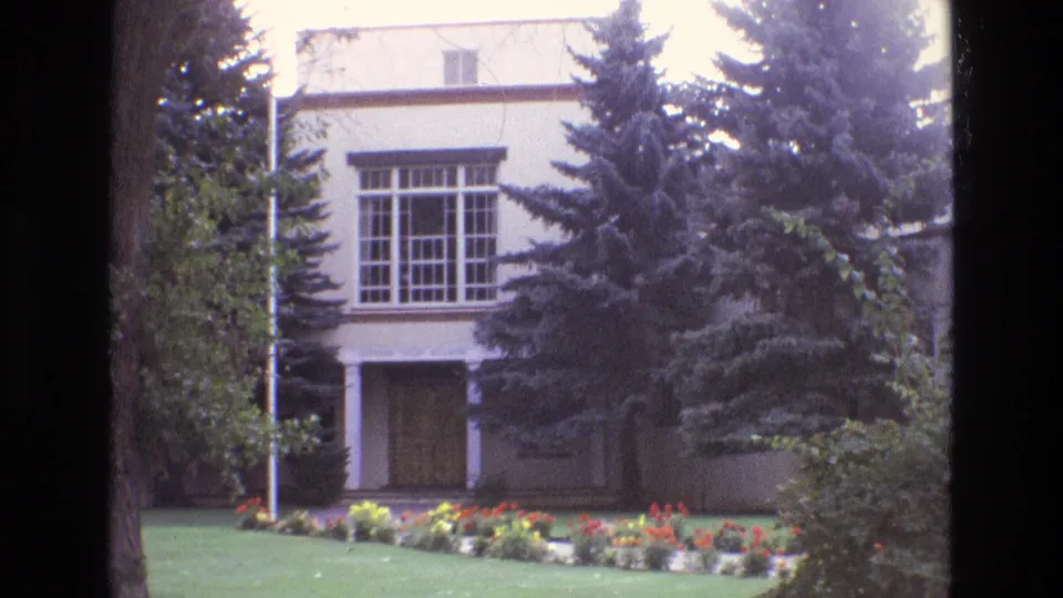 1973 New Mexico 8mm frame with a white two-story building, gridded window, shadowed covered entry, evergreen trees, shrubs, lawn, and colorful flowers.