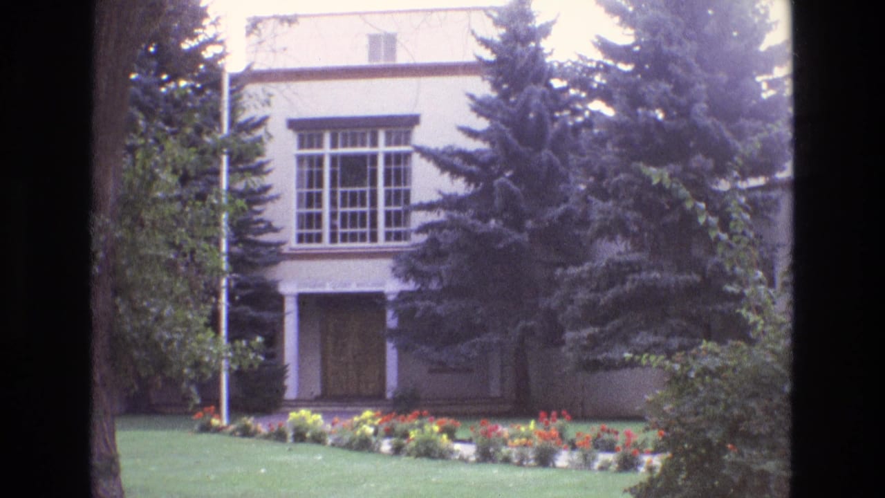 1973 New Mexico 8mm frame showing a pale stucco building facade with tall windows, evergreen trees, lawn, and a colorful flower bed.