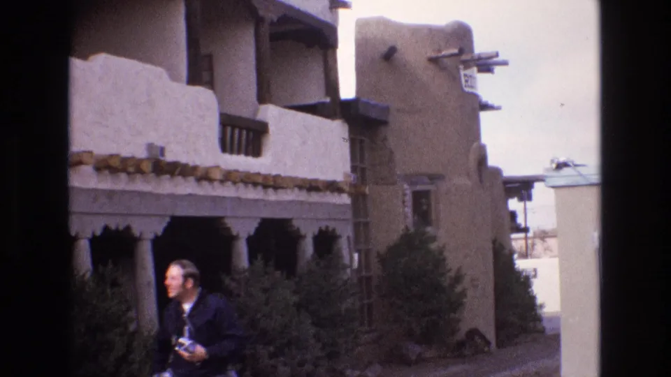 1973 New Mexico home-movie frame showing an adult man carrying a camera beside a tan adobe-style building with wooden beams and shaded arcade openings.