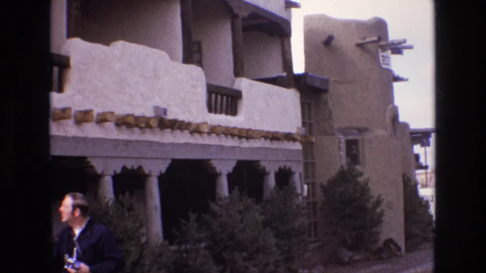 1973 New Mexico frame showing a tan adobe-style exterior with exposed beams, shaded openings, shrubs, a ladder, and an adult man partly visible at the lower left.