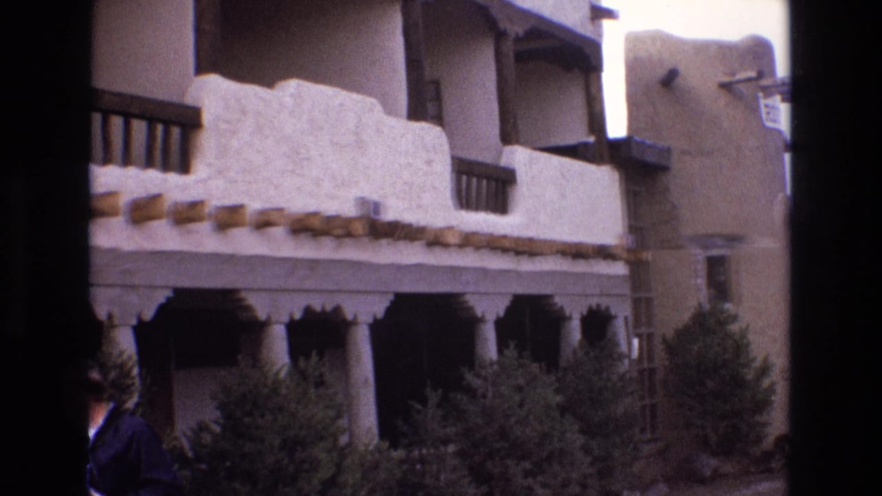 1973 New Mexico 8mm frame showing a white stucco adobe-style building with projecting wooden beams, porch columns, hedgerow planting, balcony railings, and a partially visible man at the left edge.