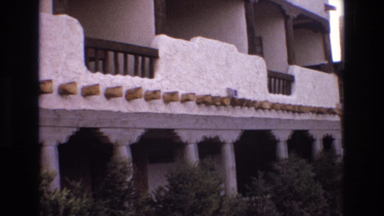 Adobe-style building facade with white stucco parapet, wooden beams, balcony railings, columns, and hedges.