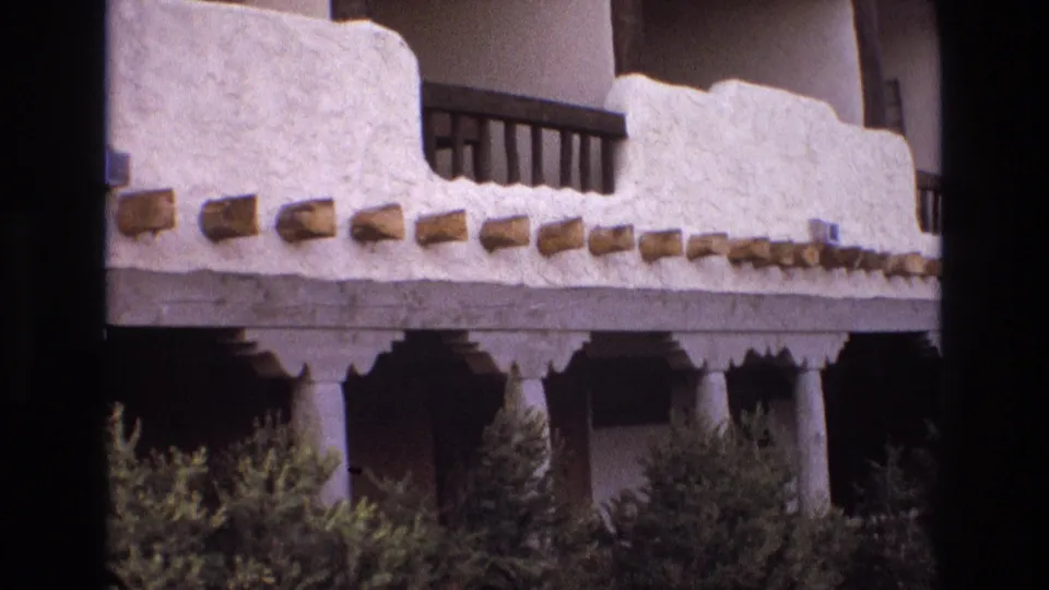 1973 New Mexico 8mm frame giving a closer view of the white stucco facade, projecting wooden beams, decorative porch trim, round columns, balcony railings, and shrubs below.