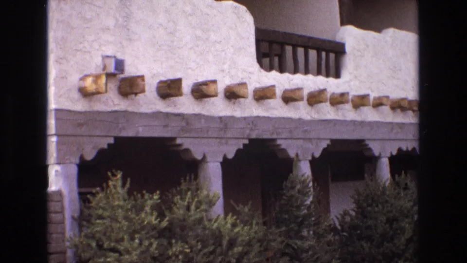 1973 New Mexico 8mm frame showing the adobe-style building facade with white textured stucco, exposed beam ends, shaded porch columns, green hedges, and a dark upper balcony railing.